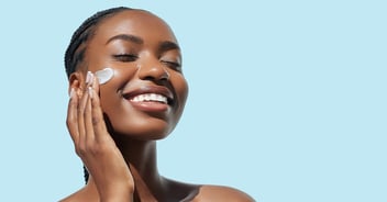 Happy young woman applying facial cream with eyes close and smiling